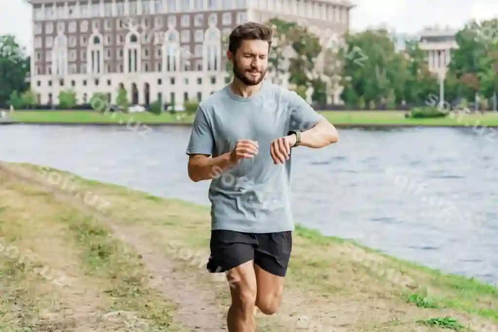 O'z attentionini qaratgan erkak yuguruvchi daryo yonida o'z akilliy soatini ko'rib chiqyotgan, o'z fitnes rivojlanishini kuzatuvchi. | Focused male runner checking his smartwatch by river tracking his fitness progress
